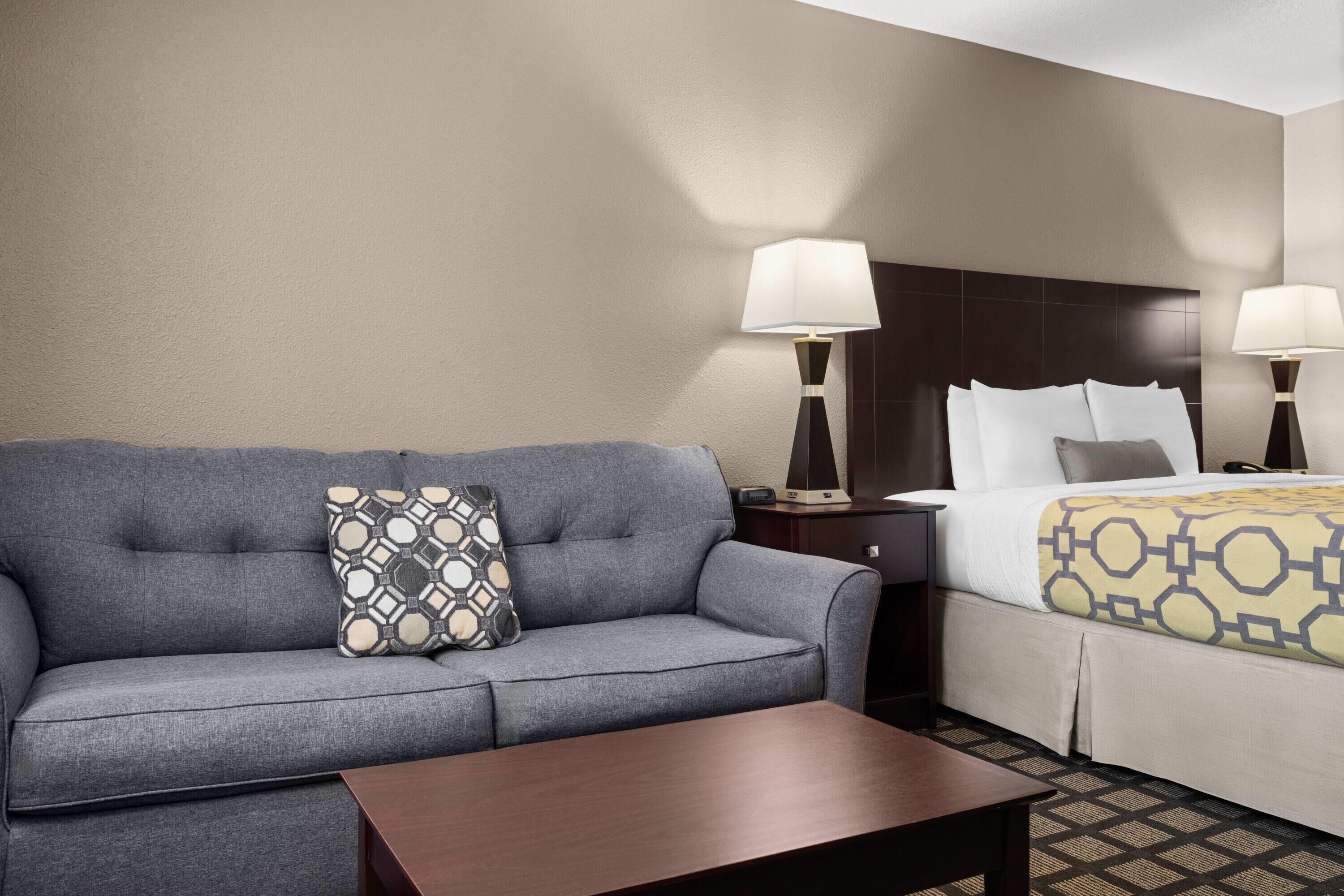 A hotel room with a gray sofa, a wooden coffee table, a bed with a patterned bedspread, two bedside lamps, and a nightstand.