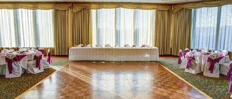 Un salón de baile con una larga mesa, sillas con cintas, una araña y amplias ventanas con cortinas.