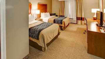 Ein Hotelzimmer mit zwei Betten, einem Schreibtisch, einem Stuhl, einer Lampe, einem Fernseher und einem Fenster mit Vorhängen.