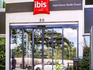 Entrada de un hotel con puerta de vidrio, un letrero rojo de 'ibis Hotels' y la dirección 'Saint Denis Stade Ouest'.