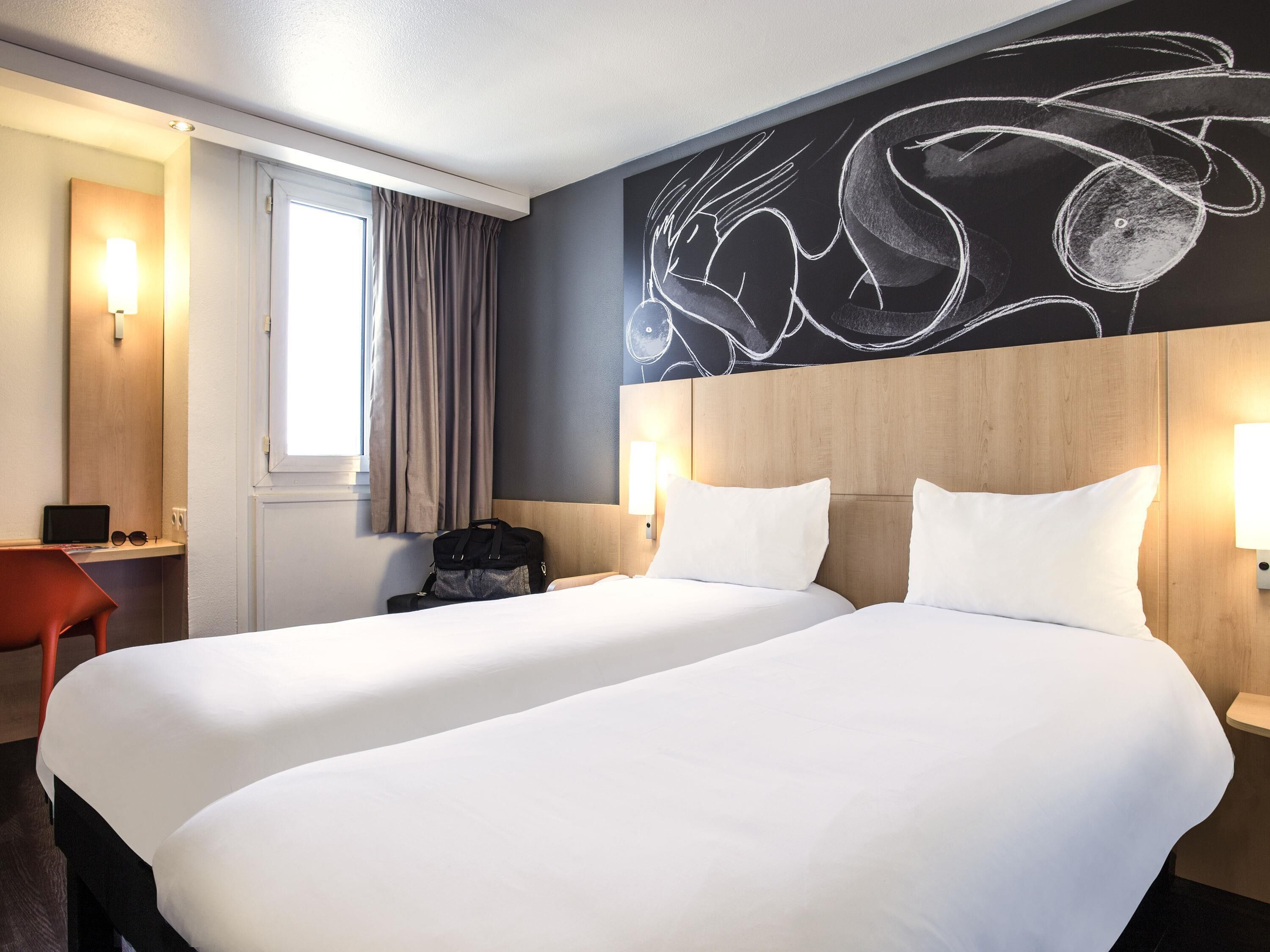 A hotel room with two beds, a desk, a chair, a wall mural, and a window with curtains.