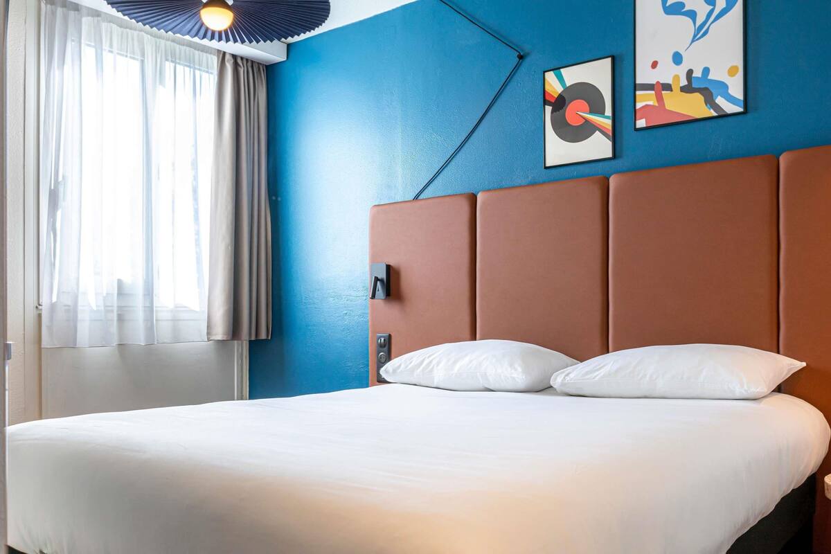 A hotel room with a bed, a window with curtains, a wall with a blue accent, and a framed picture.