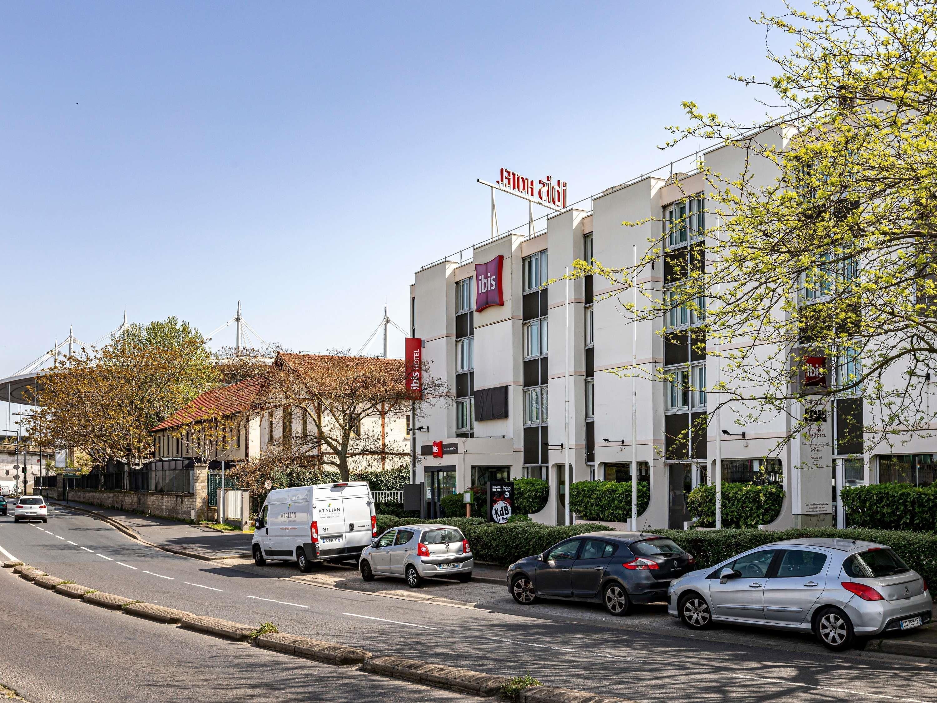 Een modern hotelgebouw met een opvallend 'ibis' bord, een parkeerplaats en een heldere lucht.