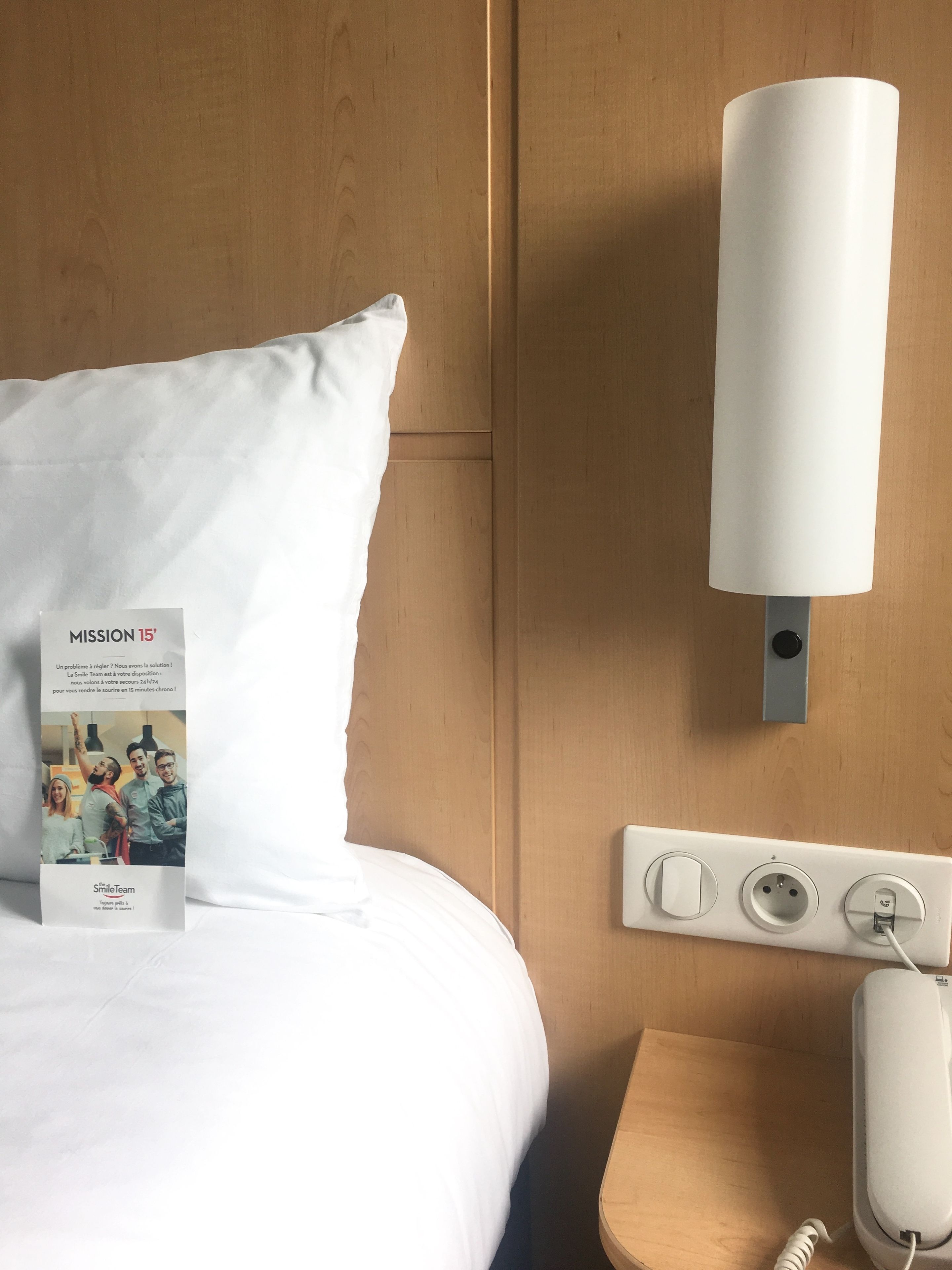 A hotel room with a neatly made bed, a bedside table with a lamp, a power outlet, and a phone.