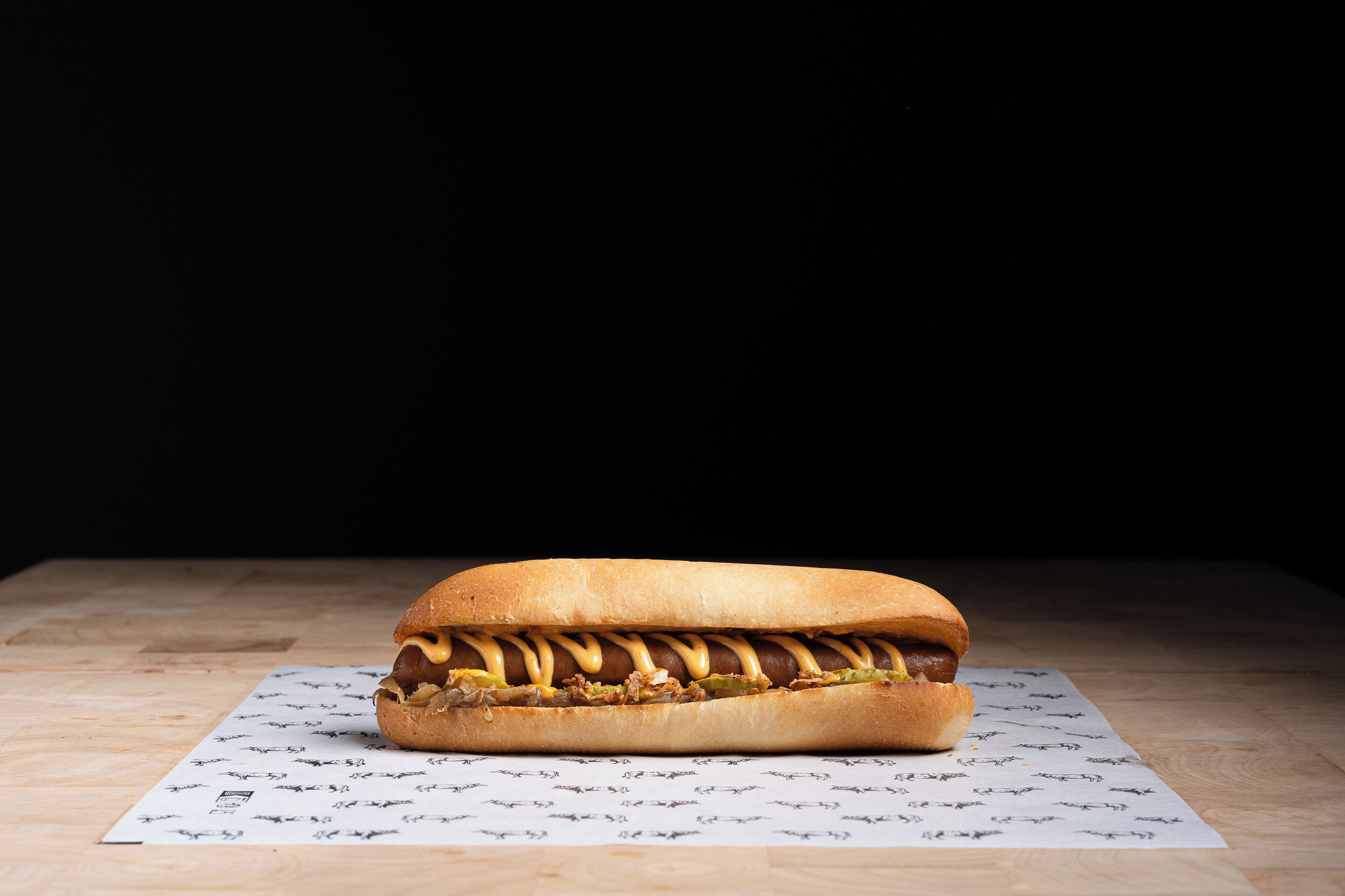 Ein Hotdog mit Senf und Gewürzgurken auf einer Holzfläche vor dunklem Hintergrund.