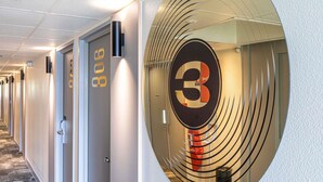 Pasillo de hotel con puertas numeradas 306 y 307, un adorno circular en la pared con el número 3, y una puerta de cristal que refleja el pasillo.