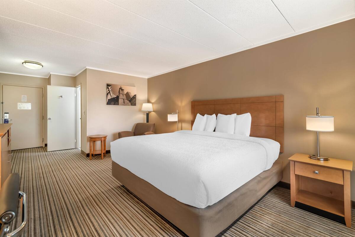 A hotel room with a large bed, two bedside tables with lamps, a desk with a chair, and a door leading to another room.