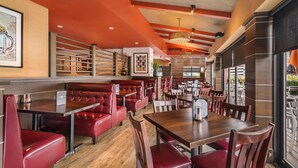 A restaurant with red booths and wooden tables, featuring a brick wall and a large window. - Best Western Plus Waltham Boston (Waltham)