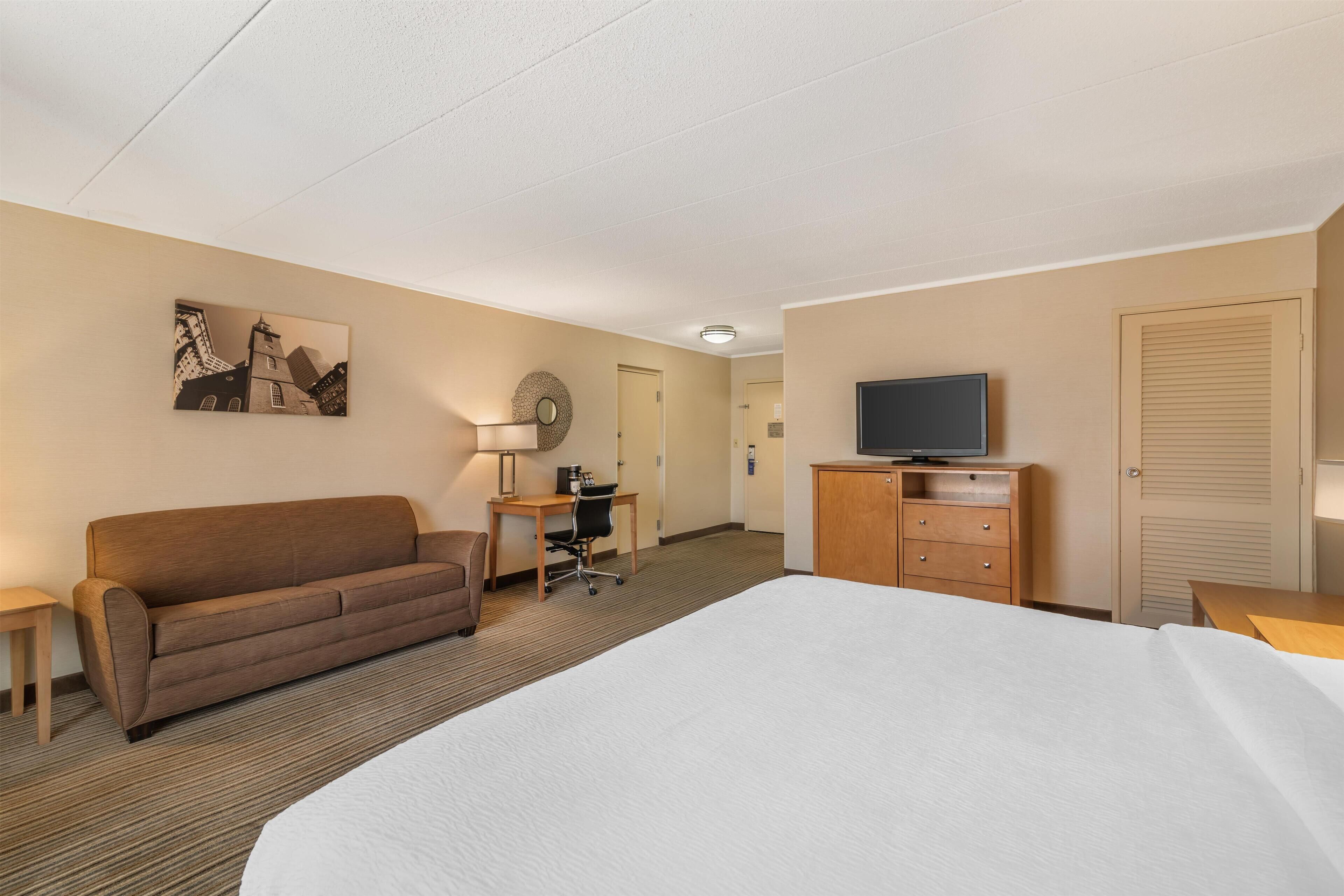Una habitación de hotel con una cama grande, un sofá, un escritorio con una silla, un televisor sobre un mueble y un cuadro en la pared.