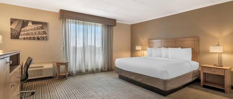 A hotel room with a large bed, a desk, a chair, a heater, and a window with curtains.