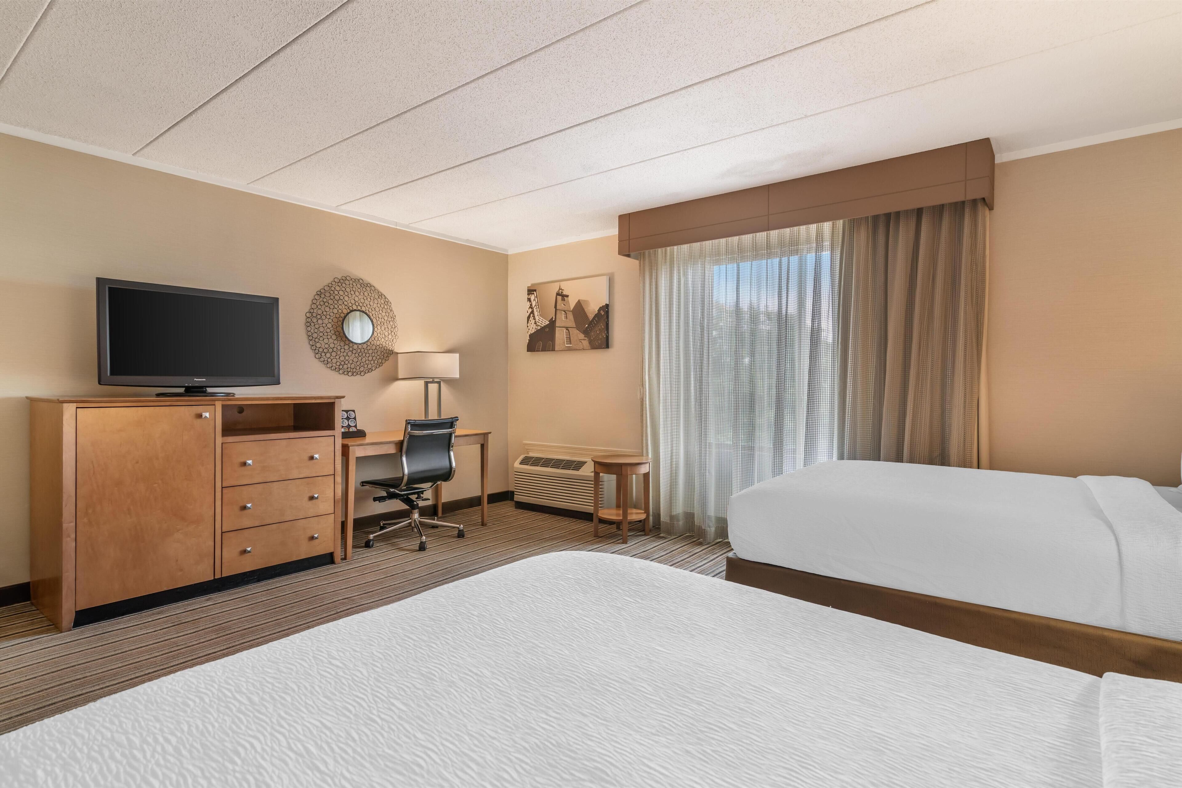A hotel room with a bed, a desk with a chair, a television, a lamp, and a window with curtains.