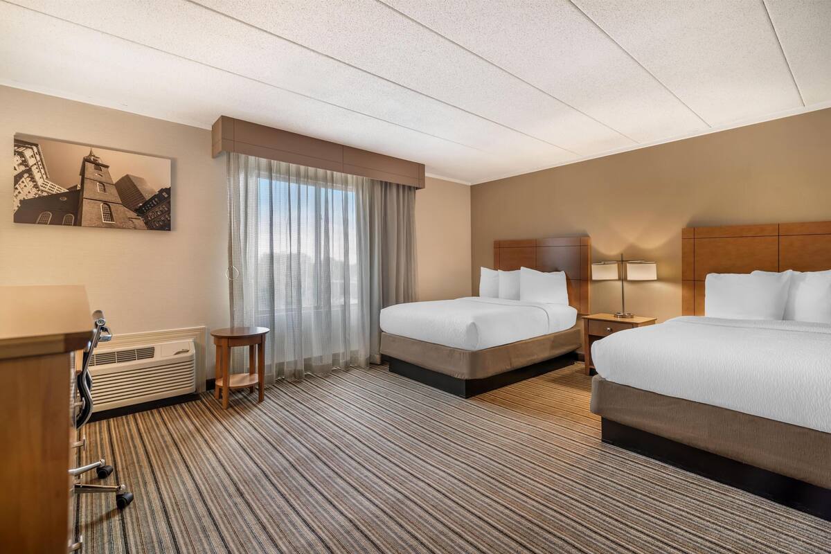 A hotel room with two beds, a desk with a chair, a small table, a heater, and a window with curtains.