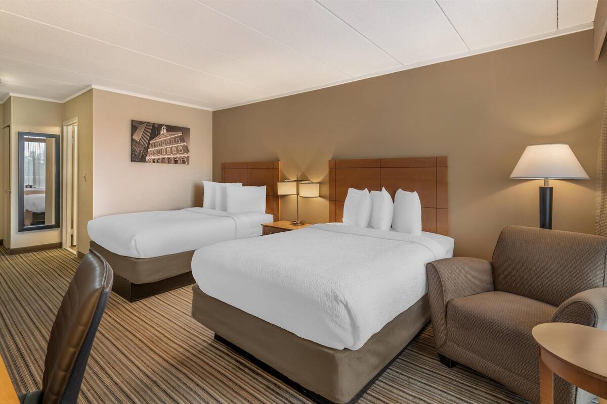 A hotel room with two beds, a chair, a lamp, and a painting on the wall.