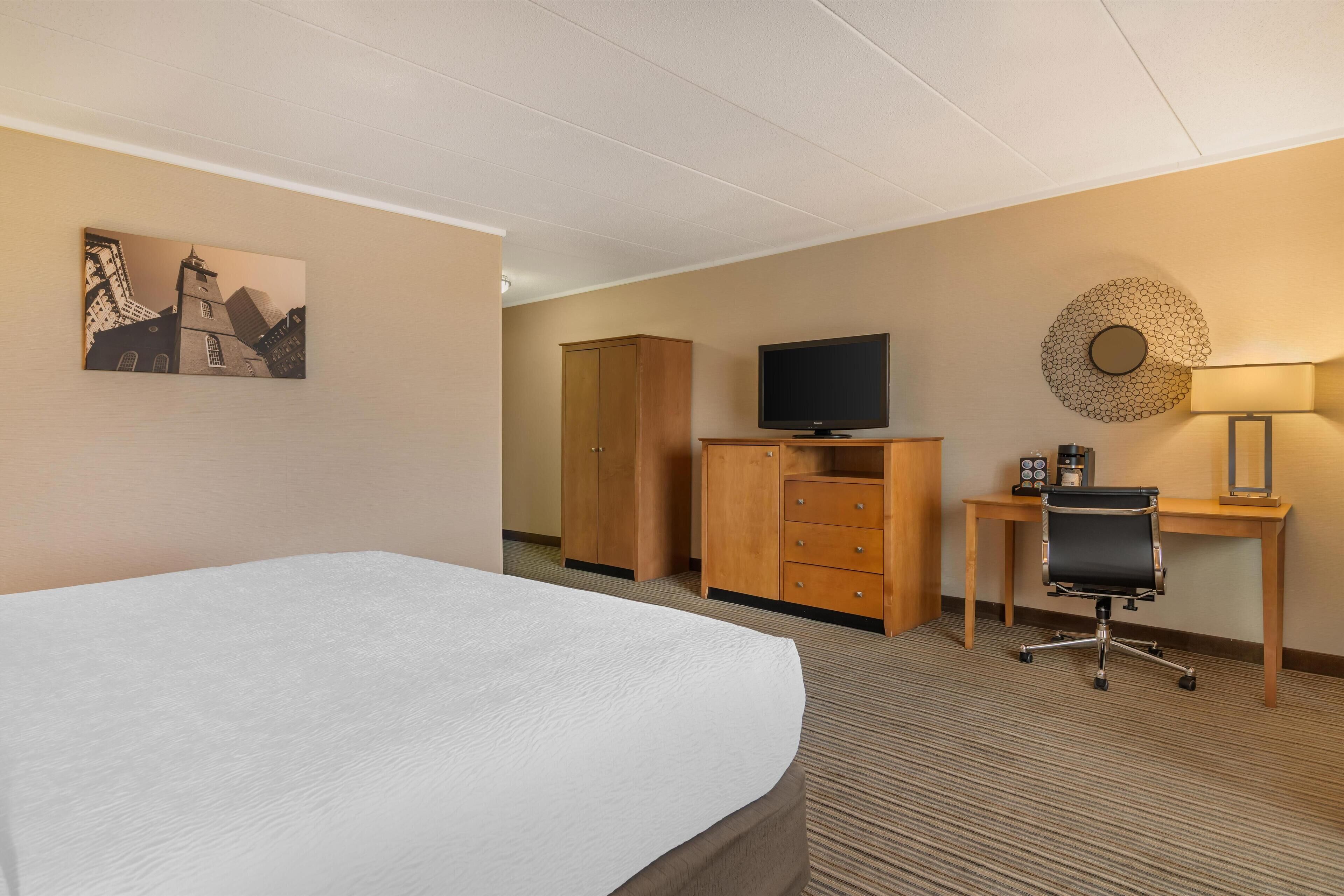 A hotel room with a bed, a desk with a chair, a television, a wardrobe, and a wall-mounted lamp.