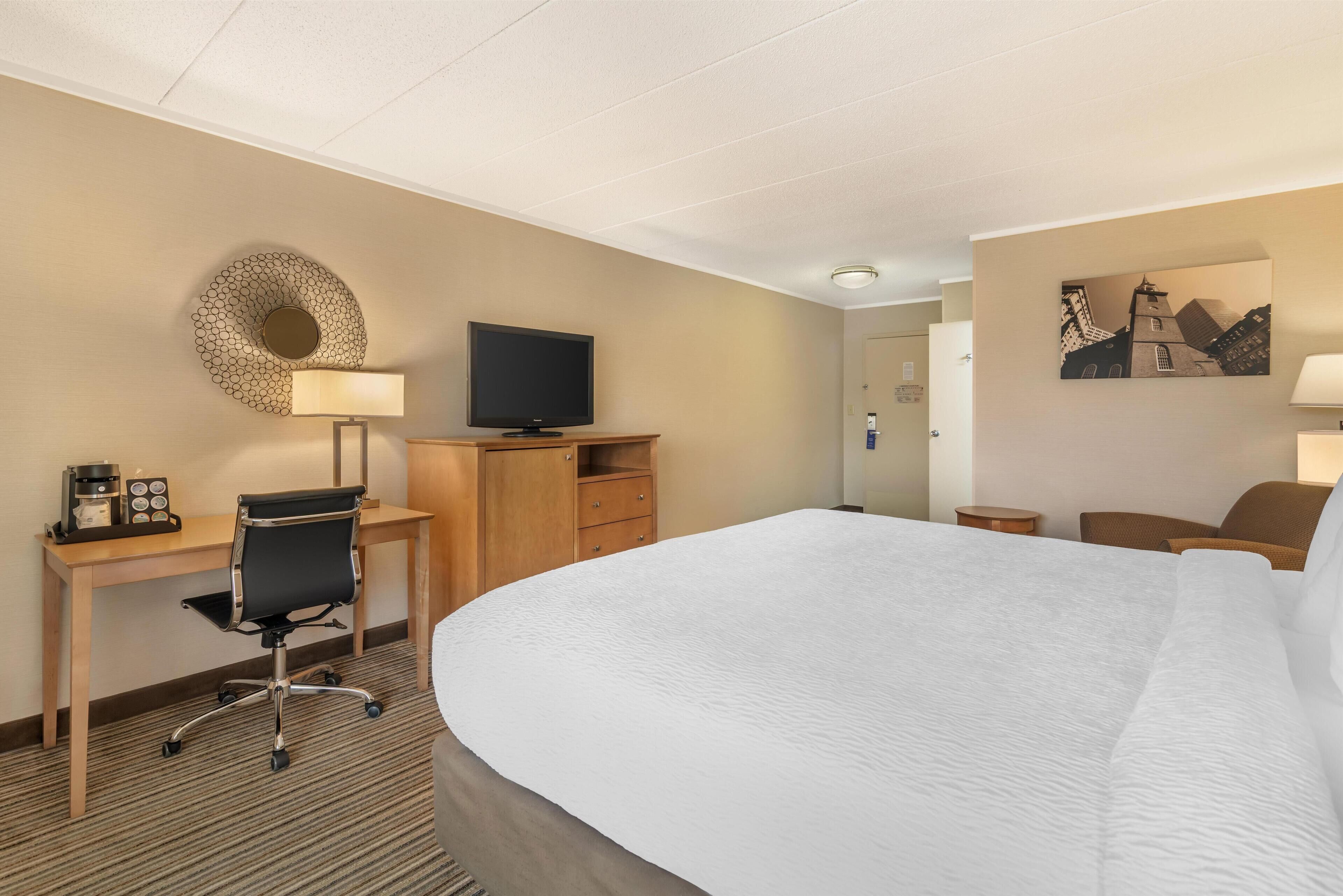 A hotel room with a large bed, a desk with a chair, a television, a coffee maker, and a wall-mounted lamp.