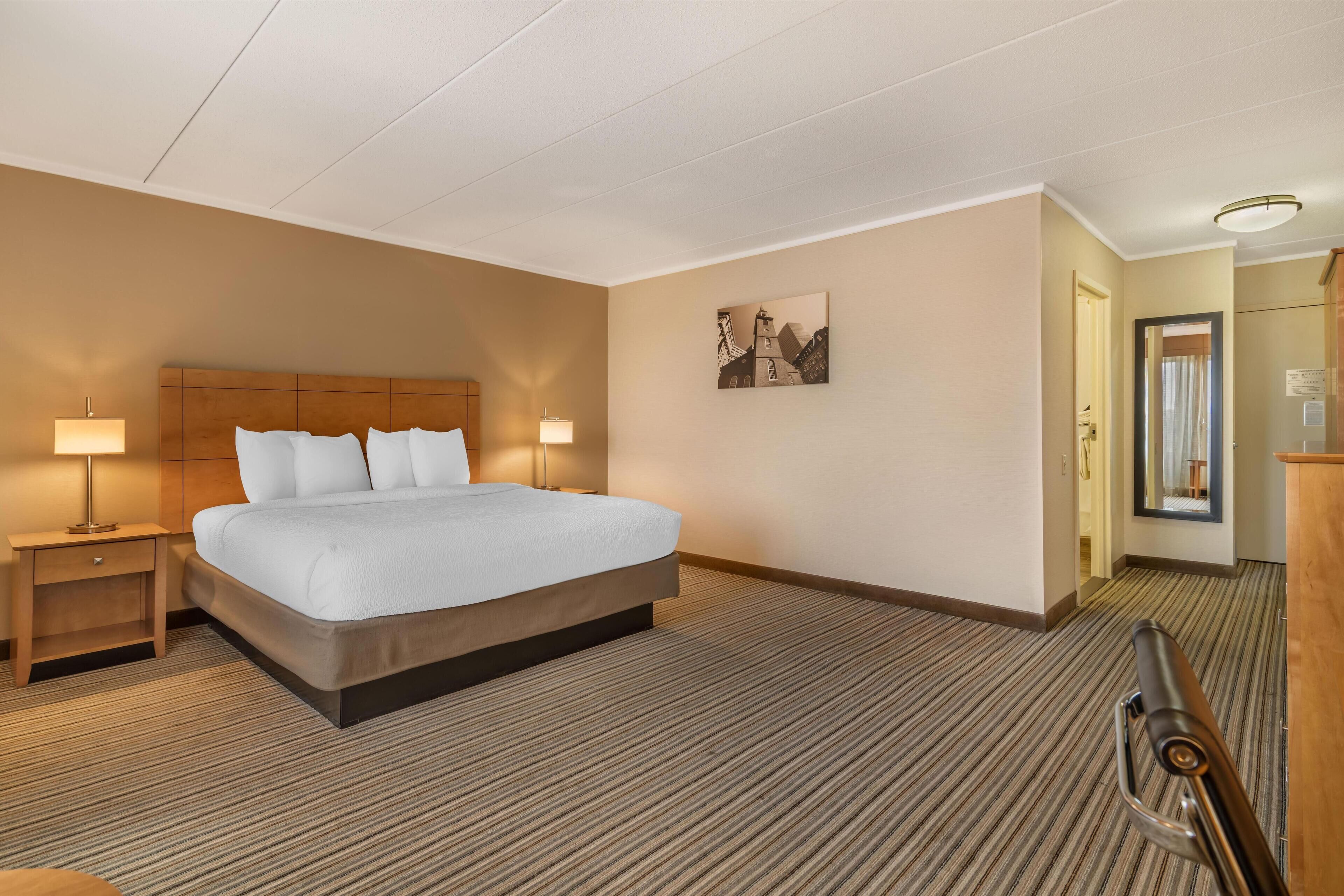 Una habitación de hotel con una cama grande, dos mesitas de noche con lámparas, piso alfombrado, un cuadro en la pared y una puerta que da a otra estancia.