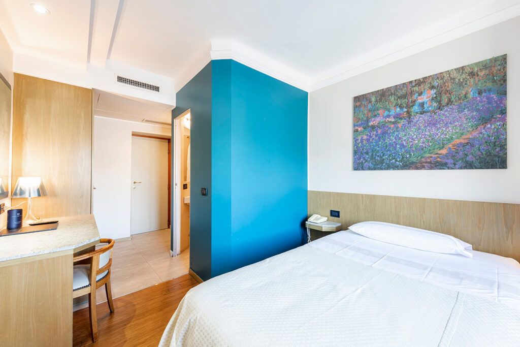 Habitación de hotel con cama, escritorio, silla, un cuadro en la pared y una puerta que da a otra habitación.