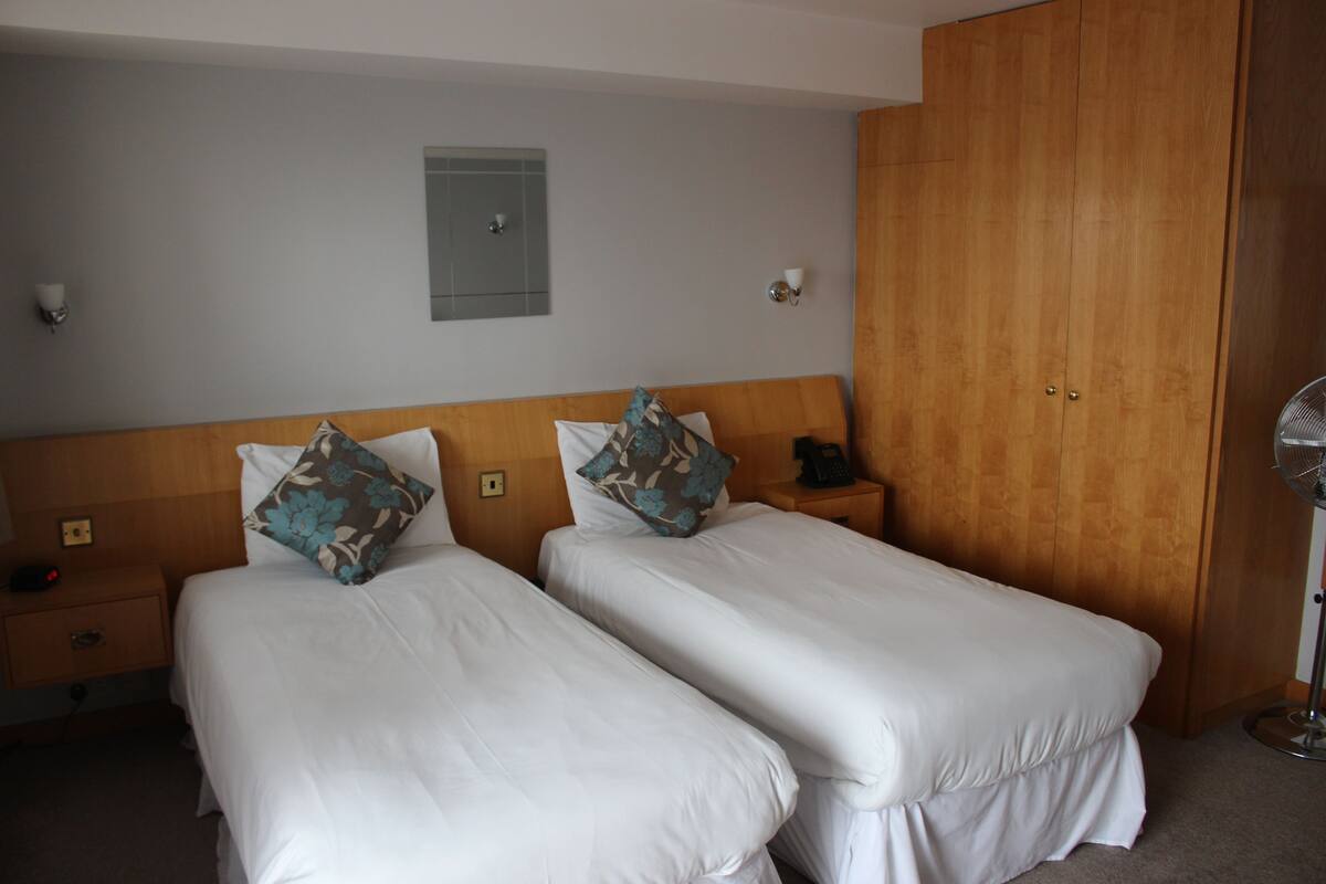 A hotel room with two single beds, a wooden wardrobe, a bedside table with a phone, and a wall-mounted light fixture.