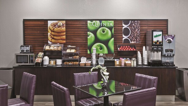 Een hotel lobby met een receptiebalie, paarse stoelen, een koffieapparaat en een muur met reclames voor eten en drinken.