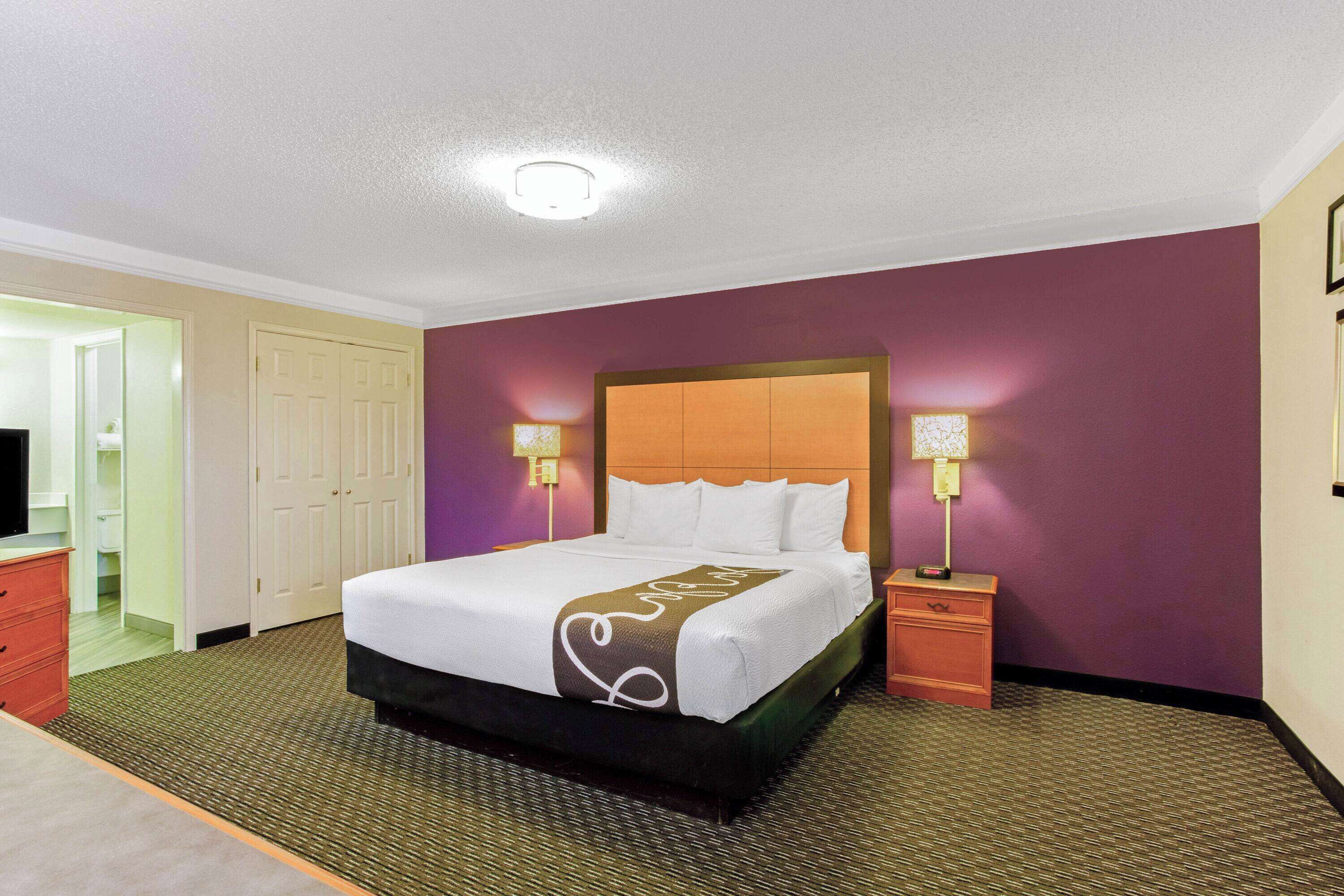 Quarto de hotel com uma cama grande, uma mesinha de cabeceira, um abajur e um guarda-roupa.