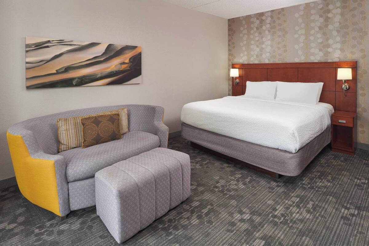 Ein Hotelzimmer mit einem großen Bett, einem grauen Sofa mit gelben Akzenten, einem kleinen Hocker, einem Gemälde an der Wand und einem gemusterten Wandpapier.