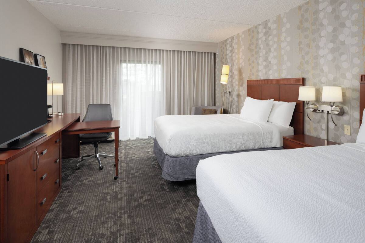 Ein Hotelzimmer mit zwei Betten, einem Schreibtisch mit Stuhl, einem Fernseher und einem großen Fenster mit Vorhängen.