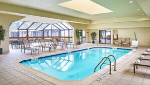 An indoor swimming pool with clear glass walls, lounge chairs, and a tiled floor.