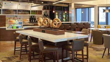 Un área de bar moderna con una barra central, taburetes, una vitrina refrigerada y un panel con la carta.