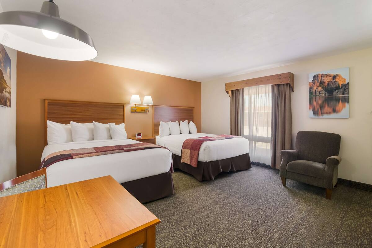 A hotel room with two beds, a wooden headboard, a desk, a chair, and a large window with curtains.