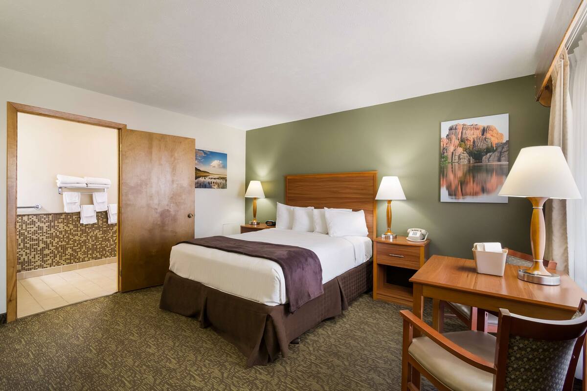 A hotel room with a bed, bedside tables, a desk, a chair, a bathroom with a shower, and a painting on the wall.