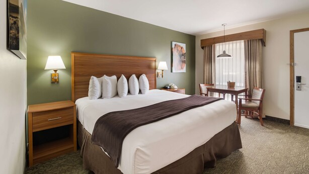 Habitación de hotel con una cama grande, un escritorio con silla, una mesita con otra silla, una ventana con cortinas y un cuadro en la pared.