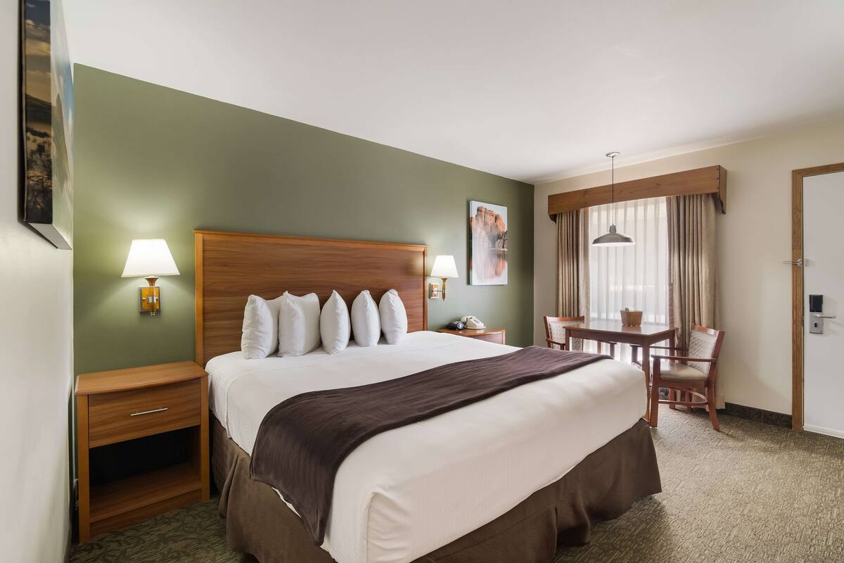 A hotel room with a large bed, a desk with a chair, a small table with a chair, a window with curtains, and a painting on the wall.