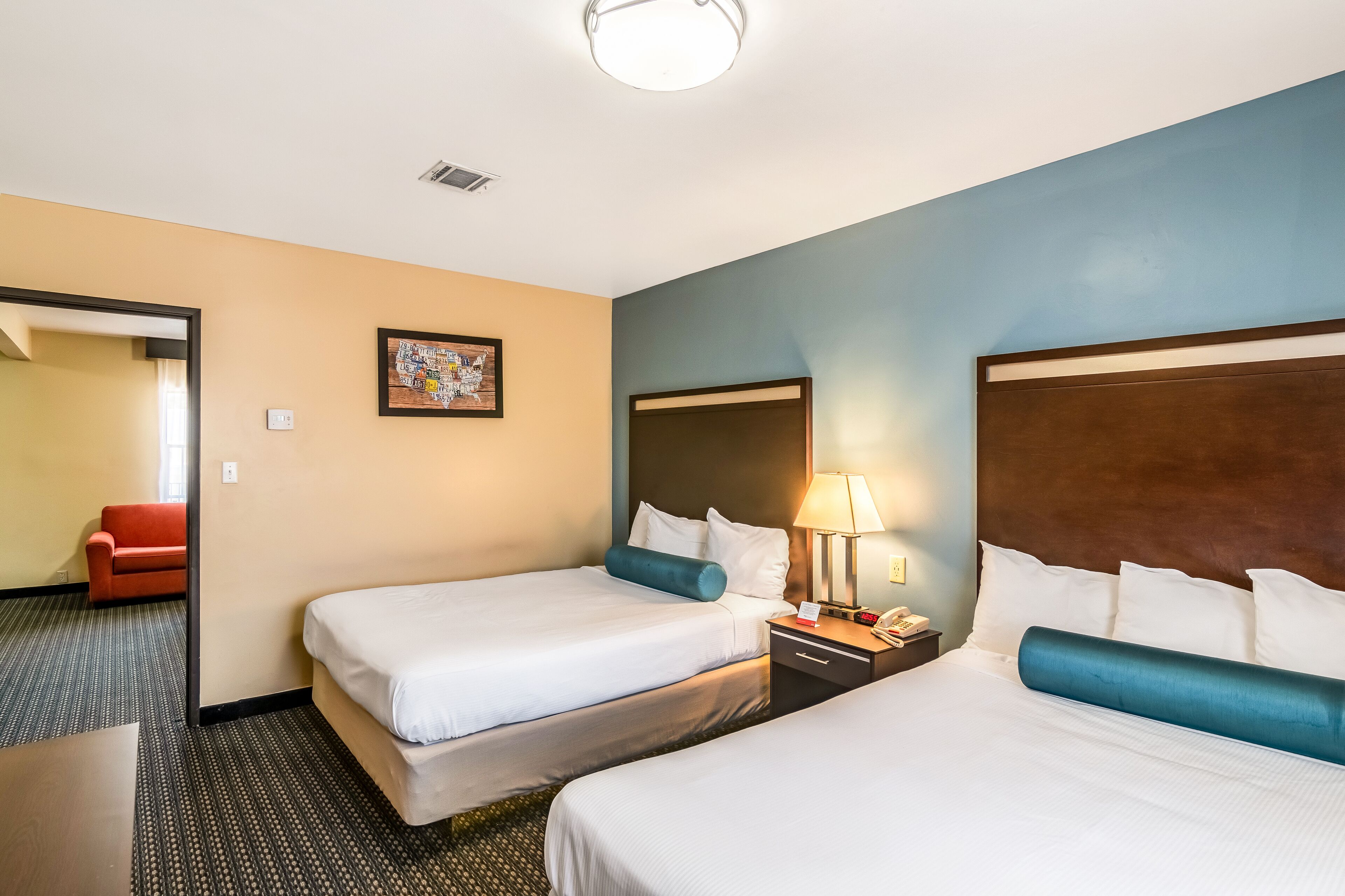 Una habitación de hotel con dos camas, una mesita de noche con lámpara, un cuadro enmarcado en la pared y una silla roja en la habitación contigua.