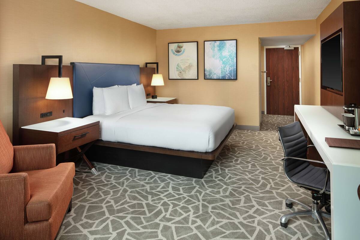 A hotel room with a bed, two bedside tables with lamps, a desk with a chair, and a bathroom door.