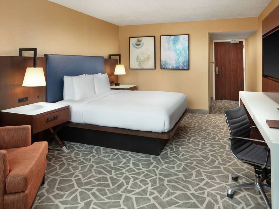 A hotel room with a bed, two bedside tables with lamps, a desk with a chair, and a bathroom door.