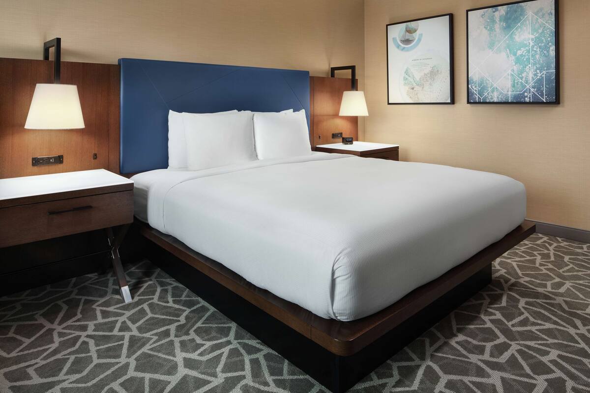 A hotel room with a large bed, two bedside tables with lamps, a wooden nightstand, and a patterned carpet.
