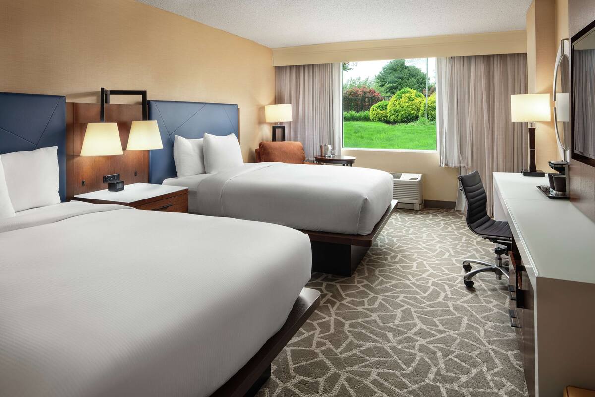A hotel room with two beds, a desk, a chair, and a view of a garden.