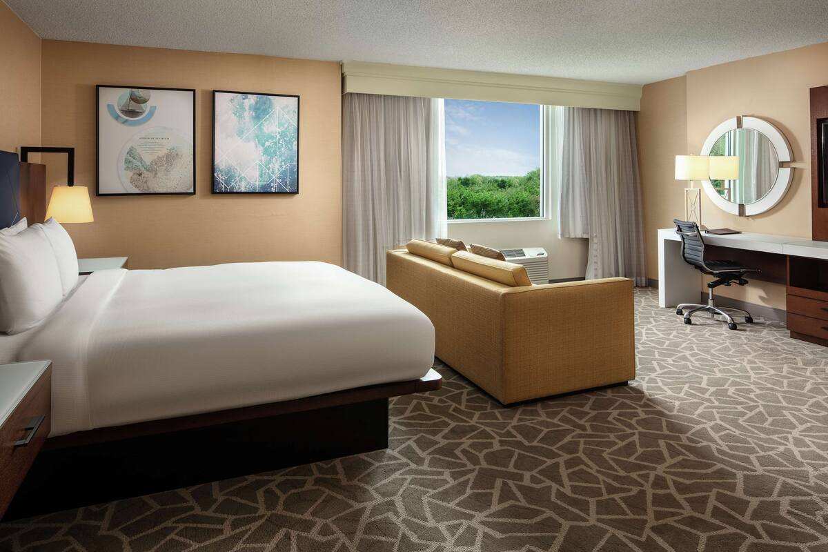 A hotel room with a large bed, a sofa, a desk with a chair, a round mirror, and two framed pictures on the wall.