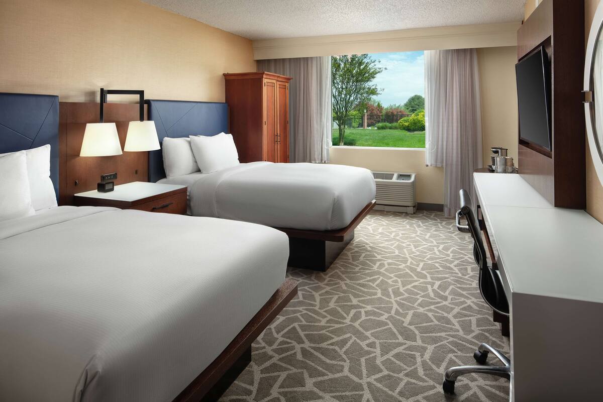 A hotel room with two beds, a desk, a TV, and a view of the outdoors.
