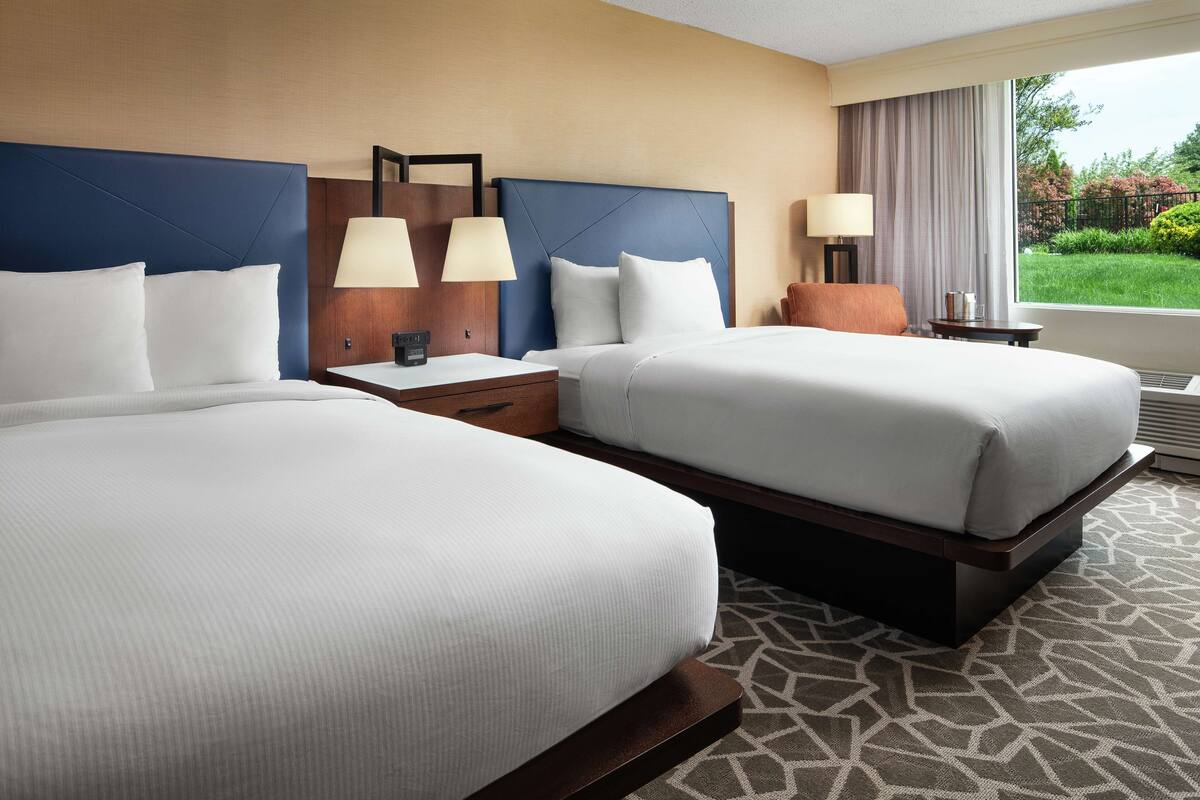 A hotel room with two beds, a nightstand with lamps, a window with a view of greenery, and a patterned carpet.
