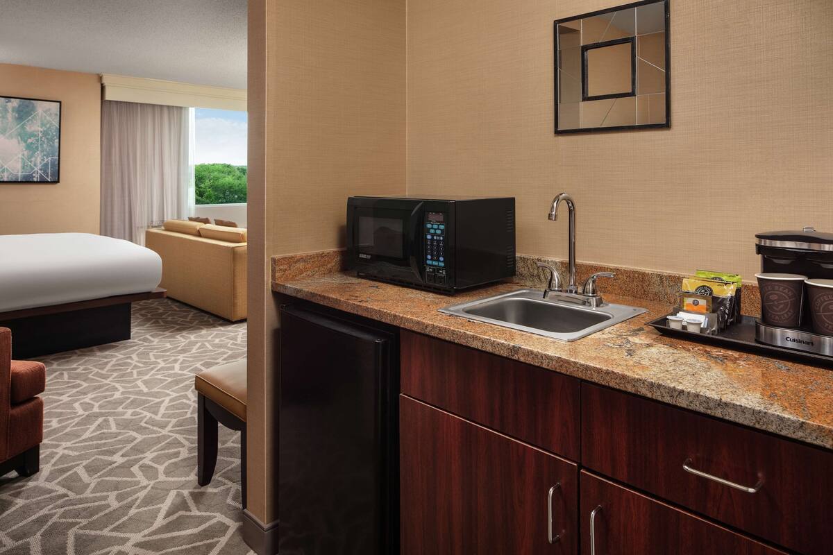A hotel room with a kitchenette, a bed, a seating area, and a view of greenery.