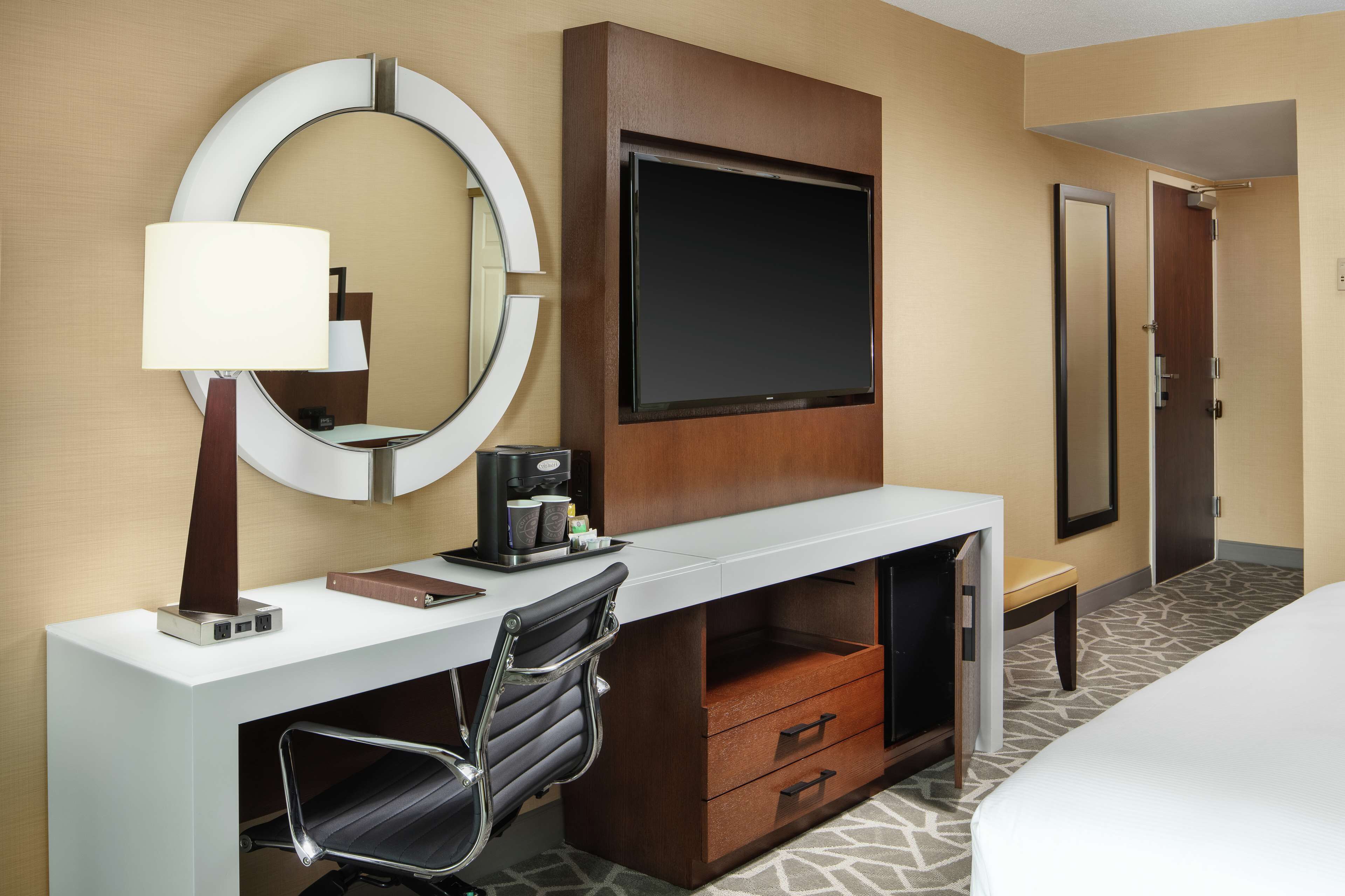 A hotel room with a white desk, a round mirror, a flat-screen TV, a coffee maker, a black office chair, a bed, and a door.