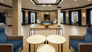 A spacious lobby with a high ceiling, a central seating area with sofas and tables, and large windows with curtains.