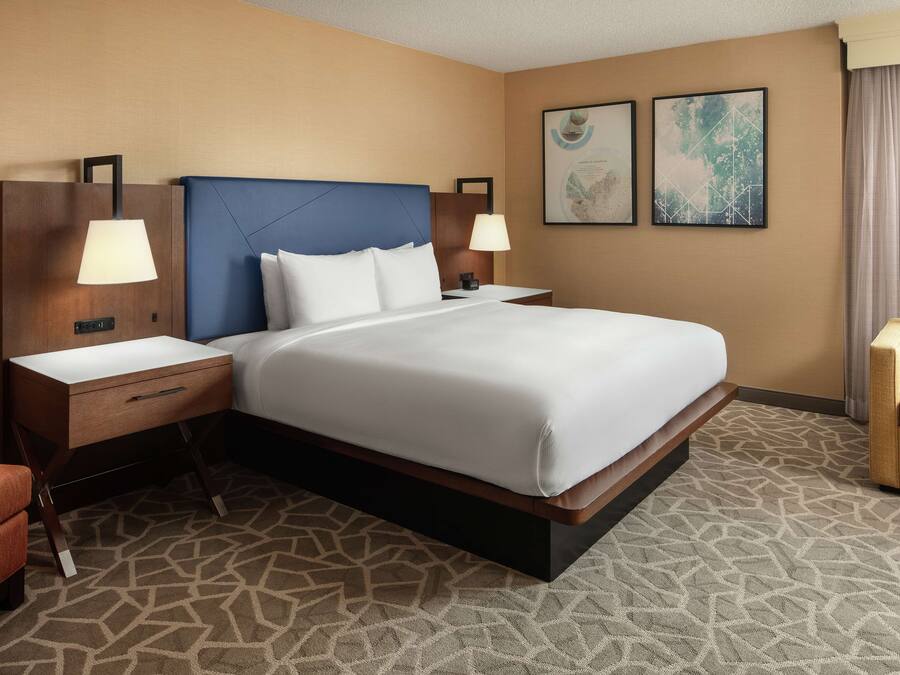 Habitación de hotel con una cama grande, dos mesitas de noche con lámparas, una silla y dos cuadros en la pared.