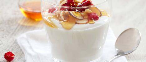 Ein Glas Joghurt mit Nüssen, Beeren und Sirup, mit einem Löffel und einer kleinen Schale Honig im Hintergrund.