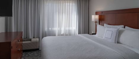 Una habitación de hotel con una cama grande, un aparador de madera, un televisor de pantalla plana y una ventana con cortinas traslúcidas.