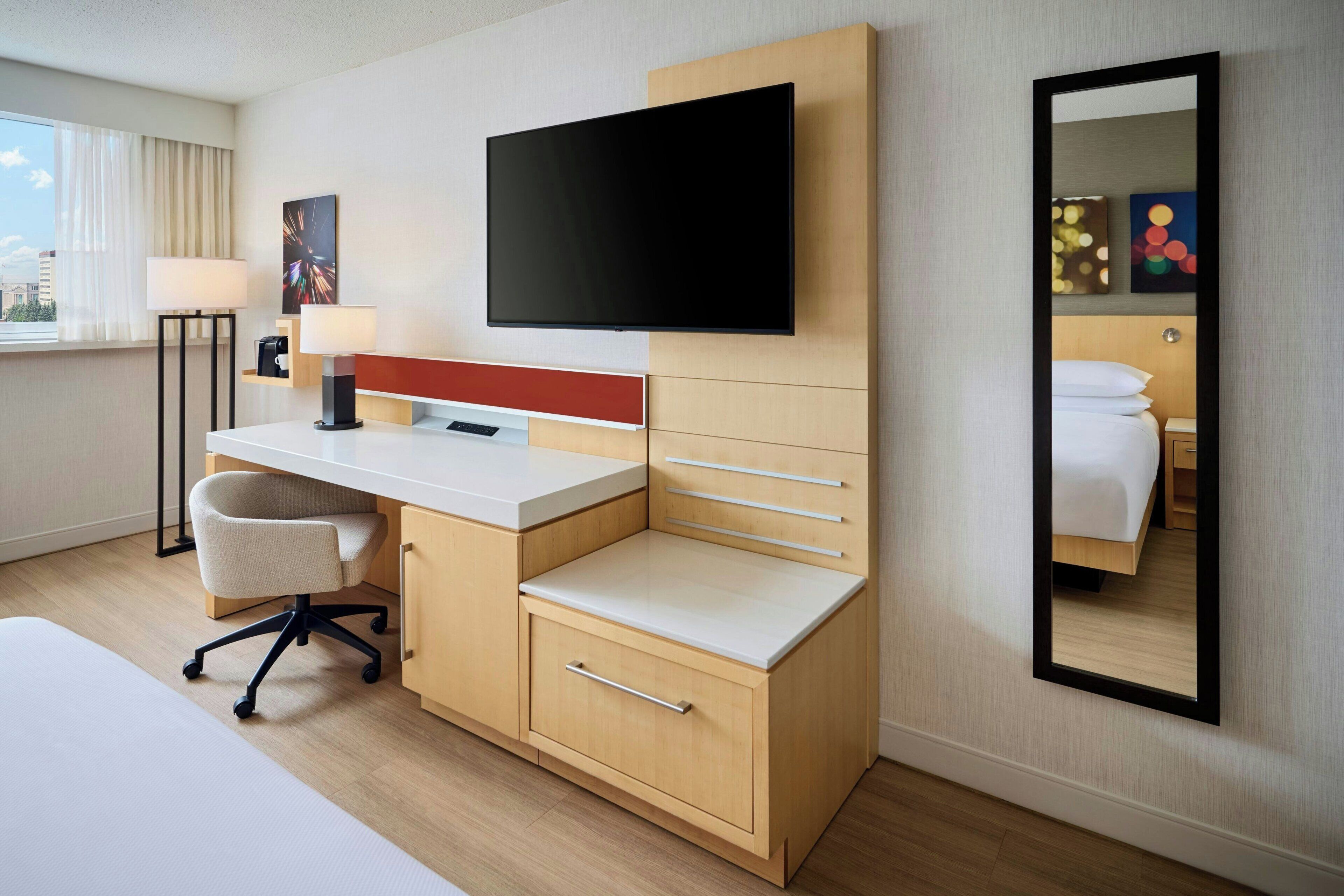 A hotel room with a bed, a desk with a chair, a flat-screen TV, a lamp, and a mirror.