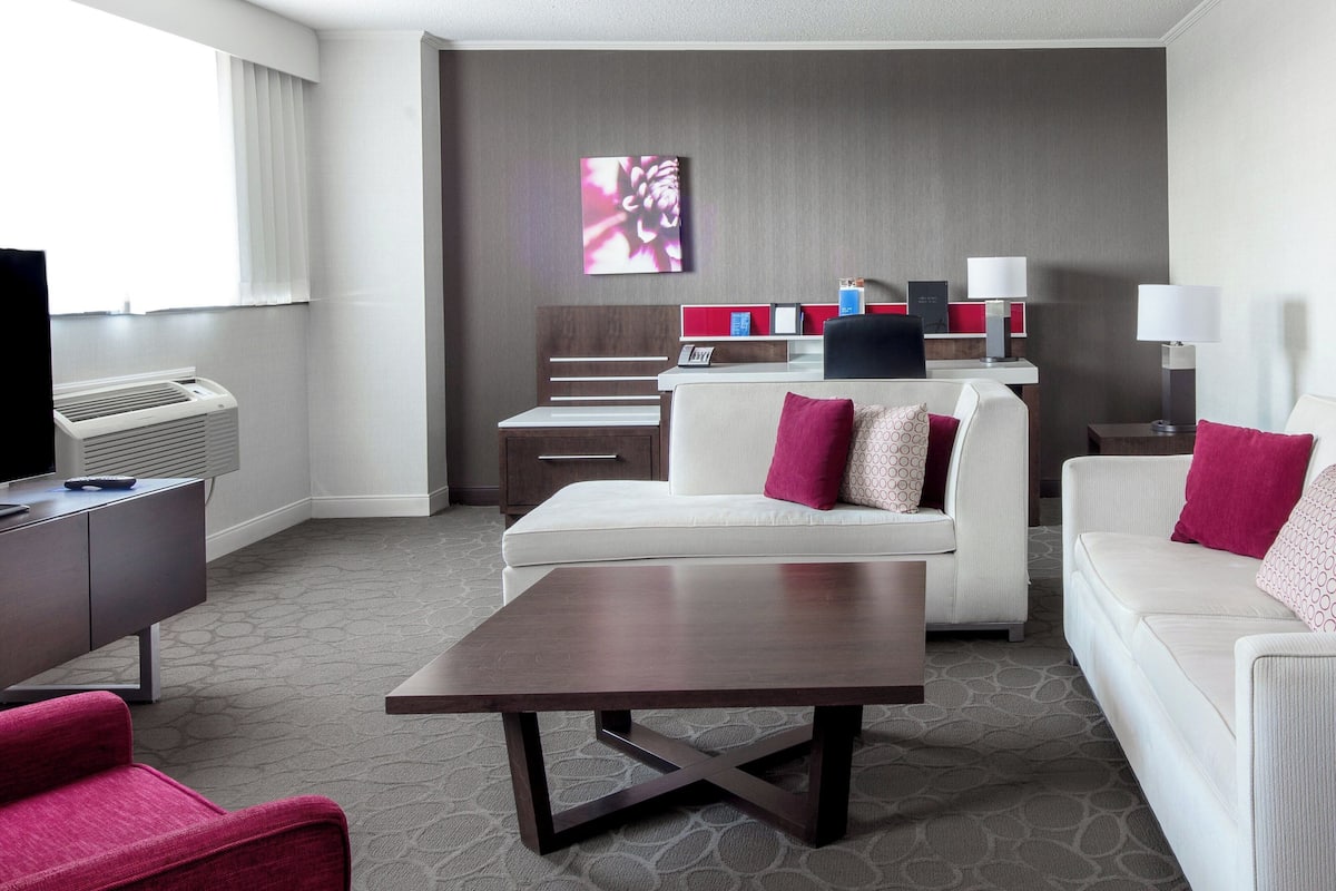 A hotel room with a sofa, a coffee table, a desk, and a TV. There is a window with blinds, a wall-mounted artwork, and a lamp on the desk.