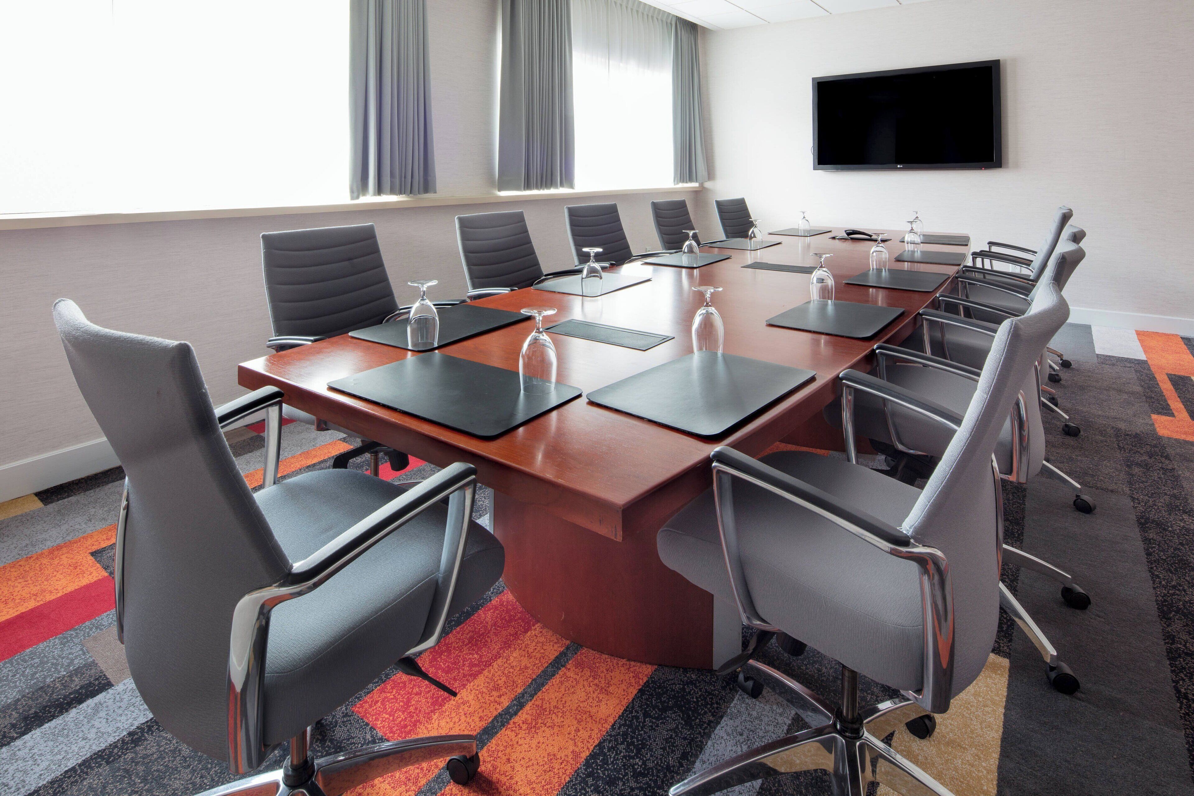 A conference room with a long table, multiple chairs, a flat-screen TV, and a window with curtains.