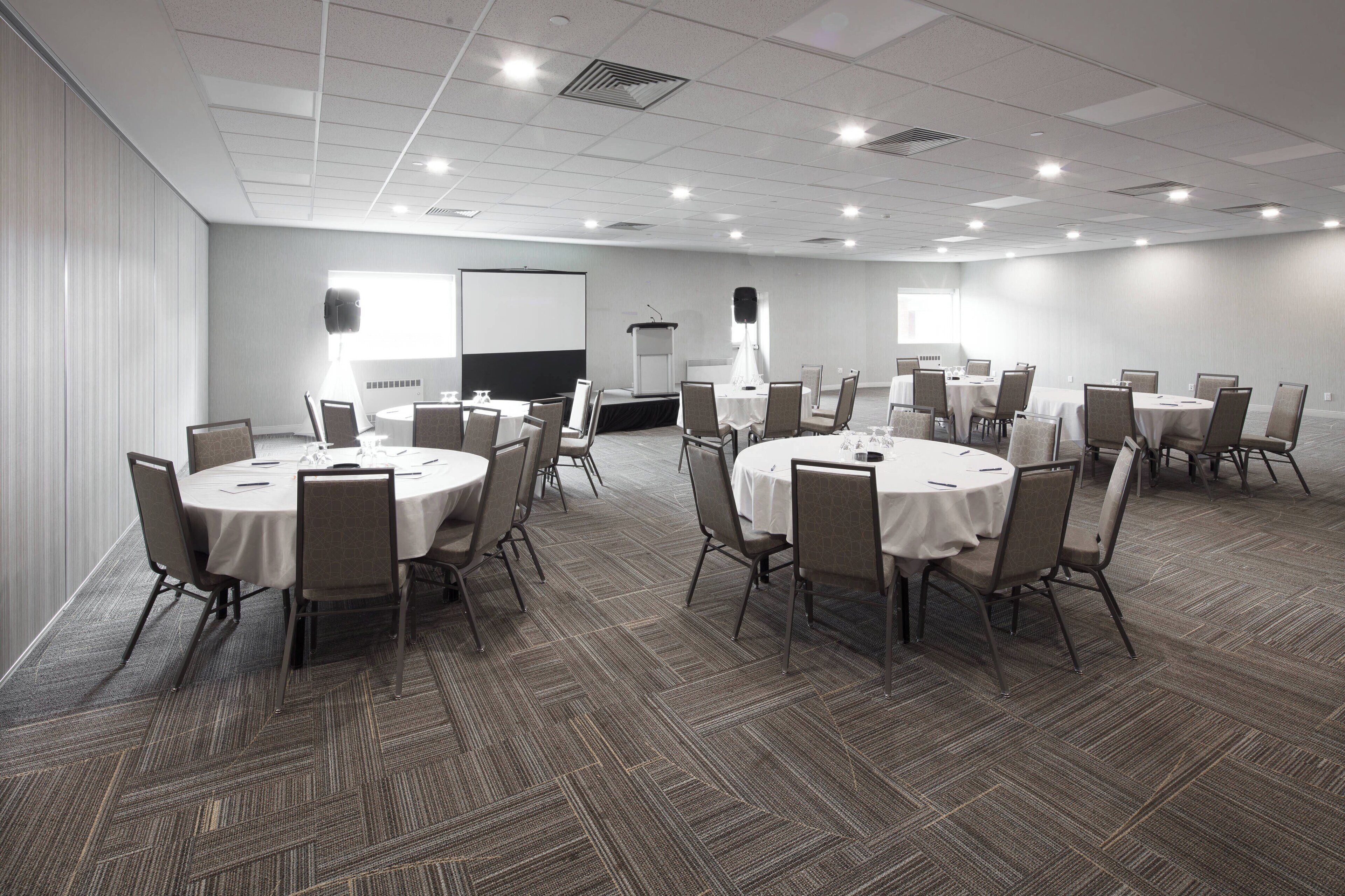 A conference room with multiple round tables, chairs, a podium, and a projector screen.